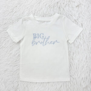 White toddler T-shirt laid on a fluffy white rug, with light blue text reading 'BIG brother' across the chest
