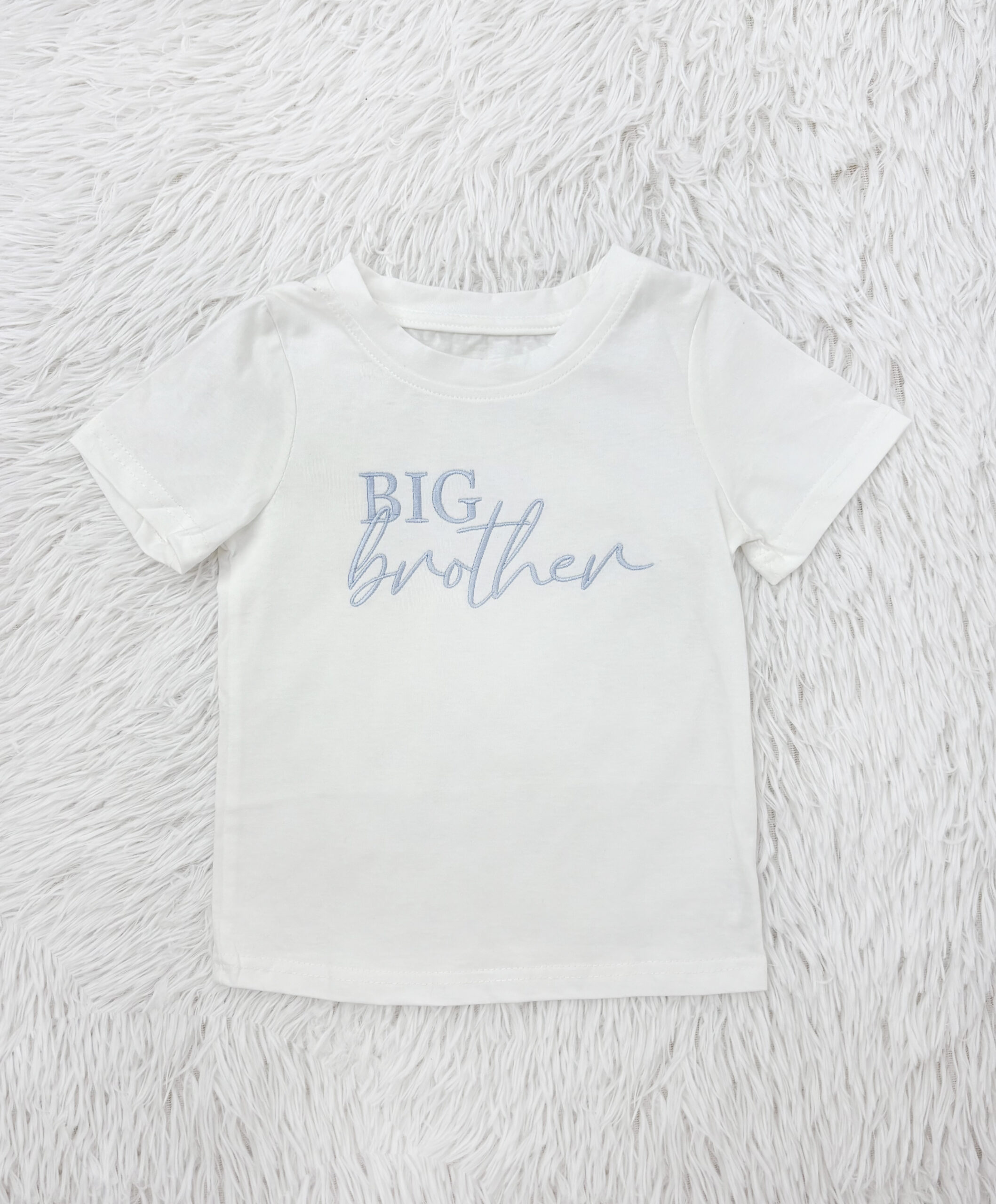 White toddler T-shirt laid on a fluffy white rug, with light blue text reading 'BIG brother' across the chest